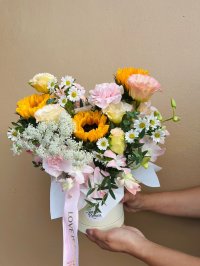 A cheerful vase bouquet with sunflowers, carnations, lisianthuses, daisies, and orchids, tied with a pink ribbon that reads “Love is Eternal.”