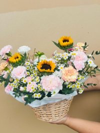 A wicker basket filled with sunflowers, pink and peach carnations, and chamomile flowers.