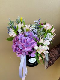 A black hat box arrangement featuring purple hydrangeas, lilac stock, pink alstroemeria, and veronica, tied with a soft lilac ribbon.
