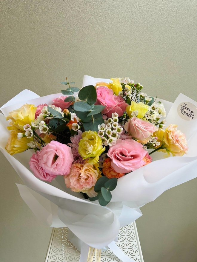 Pastel bouquet with pink roses, yellow lisianthus, peach carnations, white waxflowers, and eucalyptus wrapped in white translucent paper
