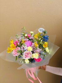 A bright bouquet of pink roses, yellow carnations, purple chrysanthemums, and blue accents wrapped in translucent paper.