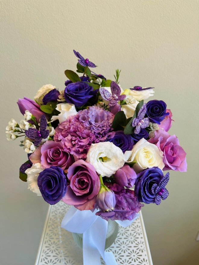Bouquet of lavender and violet roses, lisianthus, tulips, white blooms, and sparkling purple butterfly accents with a light blue ribbon
