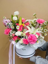 A white hat box bouquet with pink carnations, white lilies, roses, lisianthuses, daisies, and baby’s breath with pink ribbon.