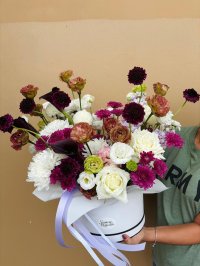 A white hat box arrangement with white roses, burgundy scabiosa, antique carnations, and purple chrysanthemums accented by calla lilies.