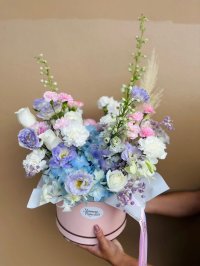 Hat box arrangement features pale lavender lisianthus, baby blue hydrangeas, delicate white roses, soft pink carnations, and airy sprigs of statice.