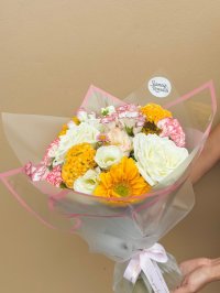 A bouquet with yellow gerberas, golden marigolds, white roses, and red-edged carnations wrapped in soft blush paper.
