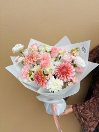 A pastel bouquet featuring dahlias, blush roses, carnations, lisianthuses, and white chrysanthemums wrapped in white paper.