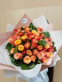 A vibrant bouquet of orange, coral, and yellow roses wrapped in blush and white paper.