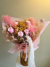 A dried floral bouquet with pink roses, golden hydrangeas, baby’s breath, and pampas in soft pink wrapping.
