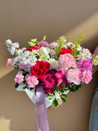 A vibrant bouquet featuring red and pink roses, lisianthuses, carnations, and hydrangeas in a white square base with purple ribbon.
