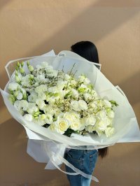 A large white bouquet with roses, lisianthus, chrysanthemums, and wax flowers wrapped in sheer white paper.