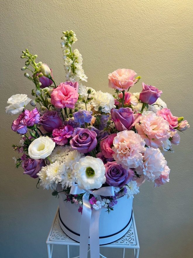 Elegant white hatbox bouquet with lilac roses, pink lisianthus, white ranunculus, and soft fillers.