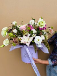 A lavender hat box with white, pink, green, and lilac flowers including lilies, lisianthuses, and roses.