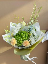 A soft green and white bouquet with peach carnations, white blooms, blue stock flowers, and green hydrangeas wrapped in olive paper.