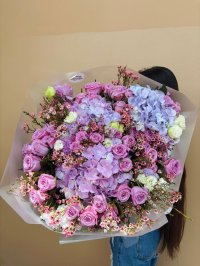 A large bouquet with lavender roses, purple hydrangeas, wax flowers, and lisianthus wrapped in pastel-toned paper.