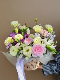 A pastel basket arrangement featuring pink roses, carnations, lisianthus, and purple hydrangeas in a woven basket.