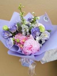  A pink peony surrounded by purple lisianthus, lavender delphinium, pale chrysanthemums, creamy anemones, and wisps of greenery, wrapped in lavender