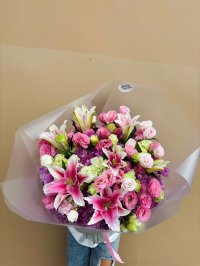 A bouquet of pink stargazer lilies, spray roses, lisianthus, and chrysanthemums wrapped in pastel paper.