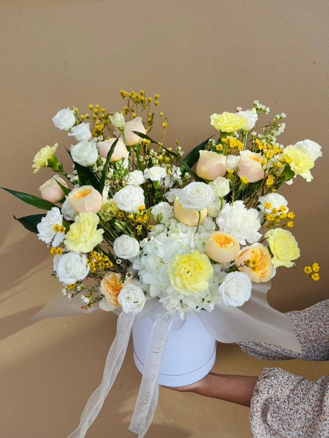 A white hatbox filled with cream and yellow roses, carnations, lisianthuses, and wax flowers.