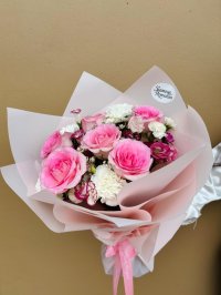 A delicate bouquet with pink roses, white carnations, lisianthus, and wax flowers wrapped in blush pink paper.