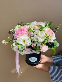 A round black hat box filled with pink roses, white daisies, lisianthus, and carnations, tied with a pink ribbon.