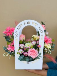 A white floral carry bag filled with pink roses, gerberas, lisianthus, and wax flowers, with “Just for You” in gold text.