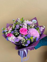 A bouquet with pink roses, lavender chrysanthemums, lilac stock, asters, and white wax flowers, wrapped in plum paper with satin ribbon.