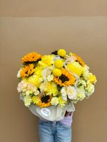 Person holding Golden Radiance Basket bouquet with orange sunflowers, yellow chrysanthemums, and white lisianthus wrapped in white ribbon against b...