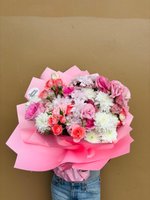 Polly bouquet featuring pink and coral roses, white chrysanthemums, and pink carnations wrapped in pink paper, held against a beige background