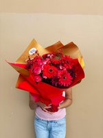 Robin bouquet featuring vibrant red gerberas, deep burgundy roses, and red chrysanthemums wrapped in red and golden paper, held by person in pink t...