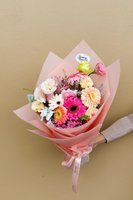 Hand-tied bouquet with pink and peach gerberas, white lisianthus, pink ranunculus, and roses wrapped in pink paper, held by a hand