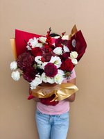 Person holding large burgundy and white flower bouquet with chrysanthemums, lisianthus, and roses wrapped in burgundy and gold paper against beige...