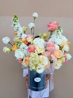 Luxury Super Peach flower arrangement in navy box featuring coral and pink roses, white lisianthus, ranunculus, and stock with pink ribbon, held by...