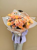 Person holding Jolene bouquet with peach roses and coral chrysanthemums wrapped in white paper with gold trim against beige background