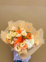 A bouquet of orange lilies, peach lisianthus, and creamy carnations with white flowers, wrapped in golden paper.