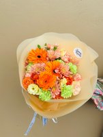 Hand-tied bouquet with orange gerberas, pink coral chrysanthemums, coral ranunculus, and light green carnations wrapped in cream paper