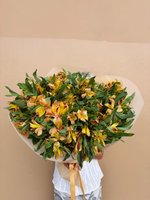 Hand-tied bouquet of yellow-orange Alstroemeria flowers with red streaks wrapped in white paper, held against a neutral background