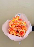 Hand-tied bouquet of 15 orange-pink gradient roses with yellow centers wrapped in pink tissue paper against neutral background