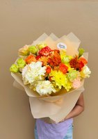 Hand-tied bouquet with orange roses, yellow chrysanthemums, and white carnations with green edges, wrapped in cream paper and held by person in pur...