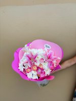 Elaine bouquet featuring pink and white striped lilies, white chrysanthemums, and pink ranunculus wrapped in bright pink paper, held against a neut...