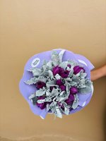 Hand holding Magic Meadow bouquet with deep purple roses surrounded by silvery-gray dusty miller foliage, wrapped in purple ribbon