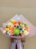 Person holding Morning Glow bouquet with orange gerberas, peach ranunculus, white lisianthus and green chrysanthemums wrapped in pink paper