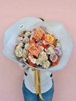 Hand-tied bouquet with peach and purple roses, white carnations, orange ranunculus and baby's breath wrapped in white paper with yellow ribbon