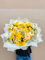 Large hand-tied bouquet of bright yellow roses and cream carnations with pink edges, wrapped in white paper, held by person in black top and jeans...