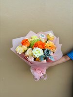 Joy Spell bouquet featuring orange ranunculus, white lisianthus, orange gerberas and green chrysanthemums wrapped in pink paper, held by hands