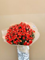 Large hand-tied bouquet featuring red roses and coral-red spray roses wrapped in white paper, held against a neutral beige background
